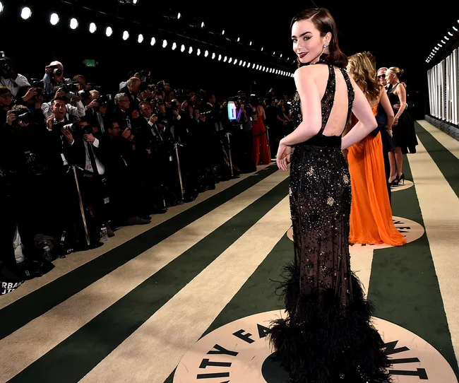 A woman in a backless black gown poses on a striped carpet at a Vanity Fair event, facing paparazzi.