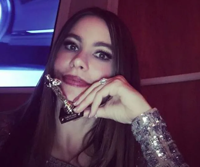 A woman with long hair holding a small trophy, wearing a sequined outfit, in a dimly lit room.