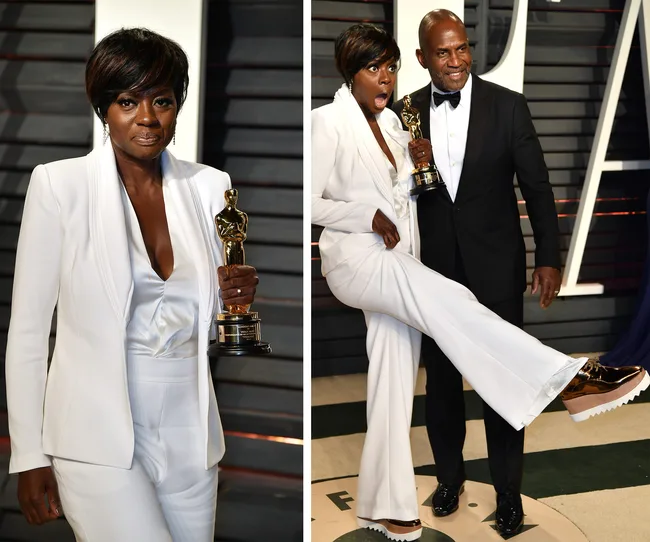 Alt text: "Person in white suit holding an Oscar, posing energetically on a red carpet with a man in a tuxedo."