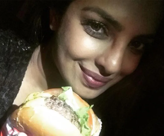 A woman smiles while holding a hamburger in dim lighting.