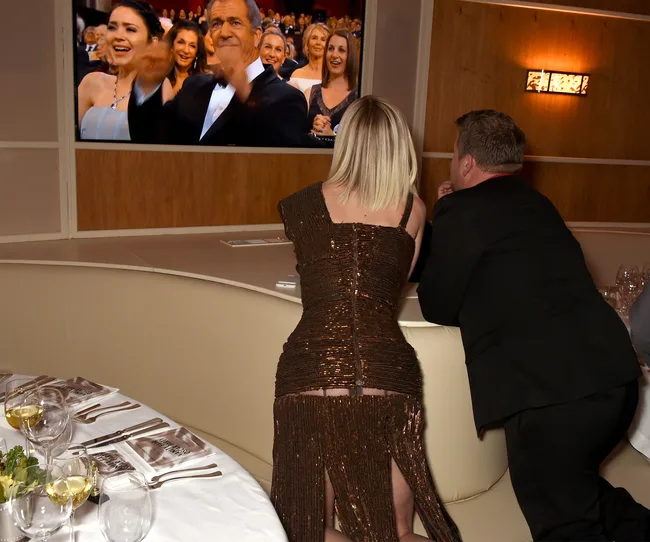 Two people in formal wear watch an awards show on a TV screen; audience visible in background.
