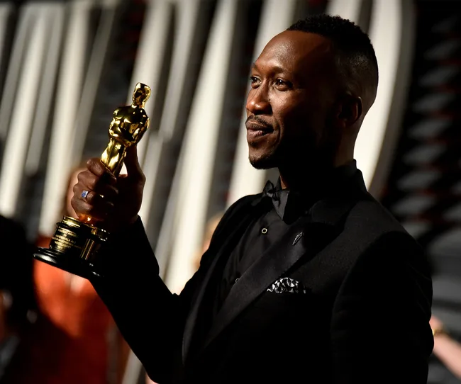 Man in a black suit holding an Oscar statue proudly, smiling at an event.