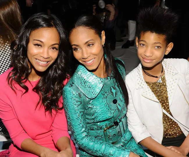 Three women sitting together smiling, one in pink, another in a turquoise outfit, and the last in a white jacket with spiked hair.