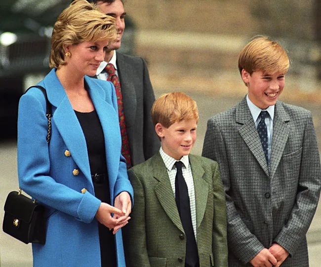 A woman in a blue coat stands with two young boys in suits, all smiling in an outdoor setting.