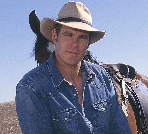 Man in a denim shirt and cowboy hat with a horse in the background under a clear blue sky.