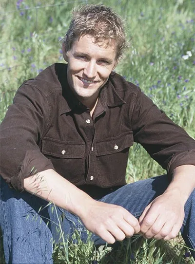 Man sitting in a grassy field, wearing a brown shirt and jeans, smiling at the camera.