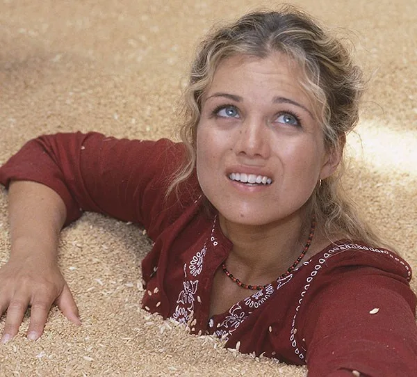 Young woman in red top partially submerged in grain, looking up with a concerned expression.