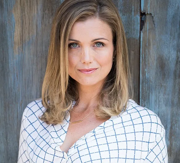 A woman with shoulder-length hair in a patterned shirt stands against a wooden backdrop.