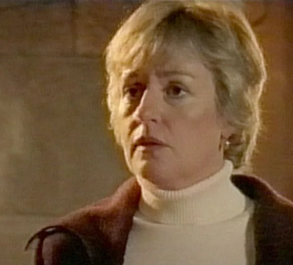 A woman with light hair wearing a white turtleneck and a dark jacket looks to the side indoors.