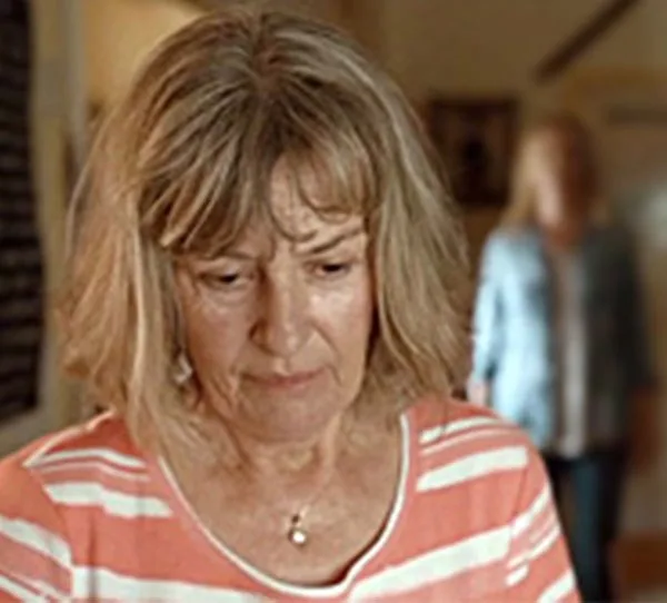 An older woman with short hair and a striped shirt looks down thoughtfully indoors.