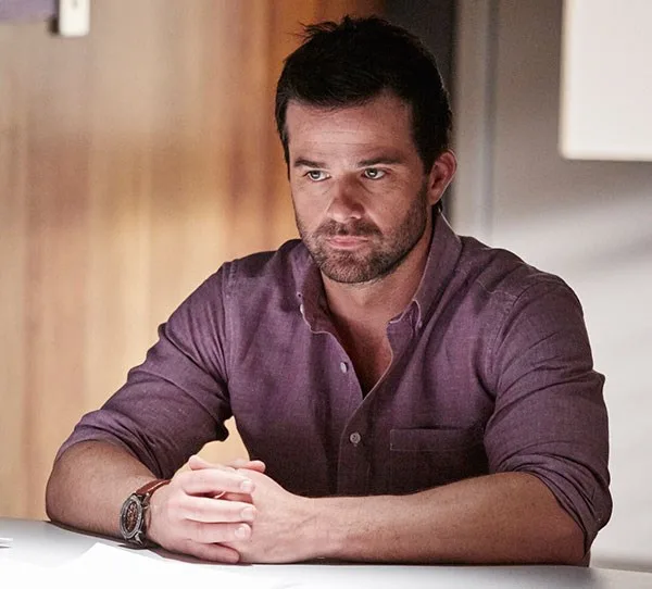 A man in a purple shirt sits at a table with hands clasped, looking serious.