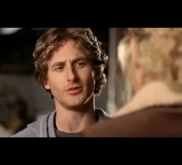 A man with curly hair talks to someone blurred in the foreground.