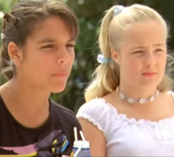 Two girls standing outdoors, one with dark hair holding a drink, the other with blonde hair in a white top and choker.