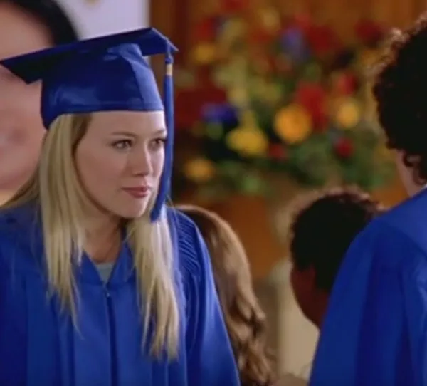 Young woman in blue graduation gown and cap, talking during a ceremony indoors with blurred background.