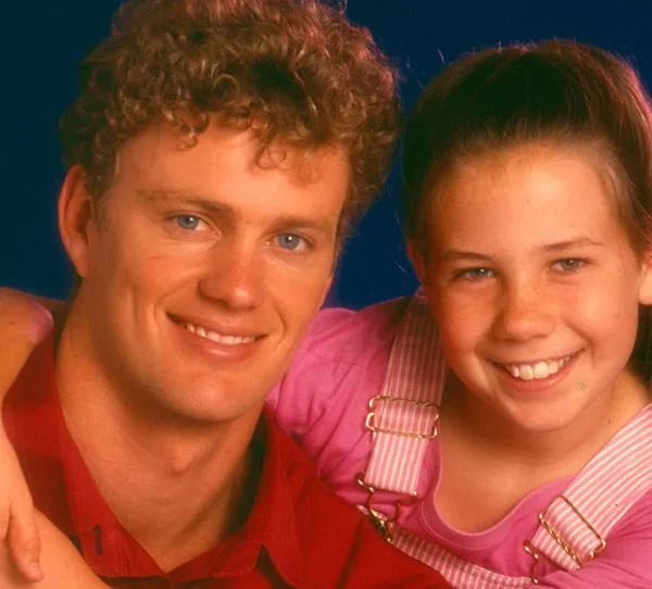 Man in red shirt and young girl in pink overalls smiling, arm around each other, against a blue background.