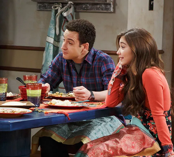 A man and woman seated at a colorful table with food, wearing aprons, in a cozy kitchen setting.