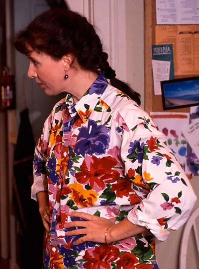 Woman in floral blouse with braided hair, standing in profile indoors, hands on hips.