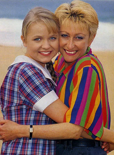 Two women smiling and embracing on a beach. One wears a plaid shirt, the other wears a colorful striped shirt.
