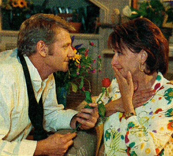 Man offers a woman a red rose, both smiling, in a warmly lit room filled with plants.