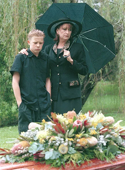 A woman and a boy dressed in black stand under an umbrella at a funeral beside a casket adorned with flowers.