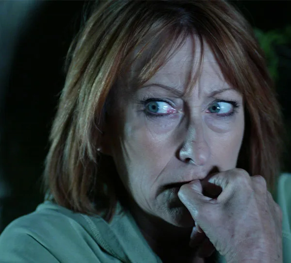 An elderly woman with a worried expression biting her knuckle, looking to the side, in a dimly lit setting.