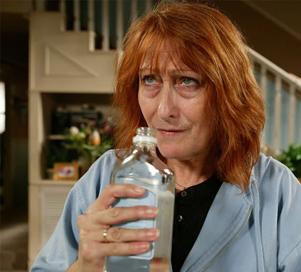 Older woman with red hair holding a clear bottle indoors, looking contemplative.