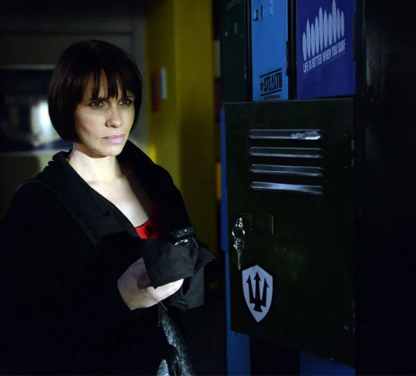 A woman stands in front of a locker, holding items, in a dimly lit hallway.