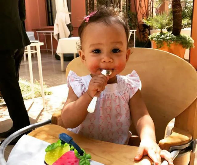A baby in a high chair, wearing a white dress, is holding a spoon in an outdoor setting.