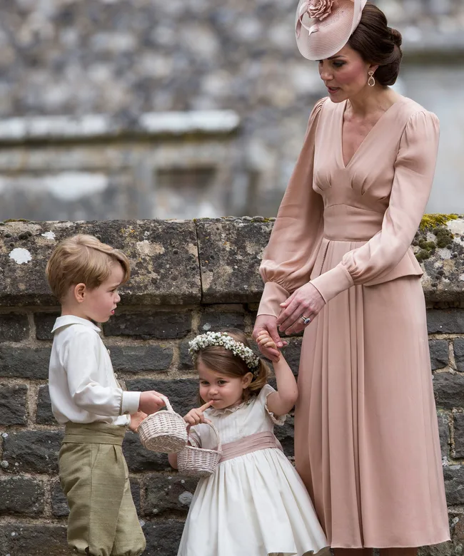 A woman in a pink dress and hat stands with two dressed children holding baskets against a stone wall.