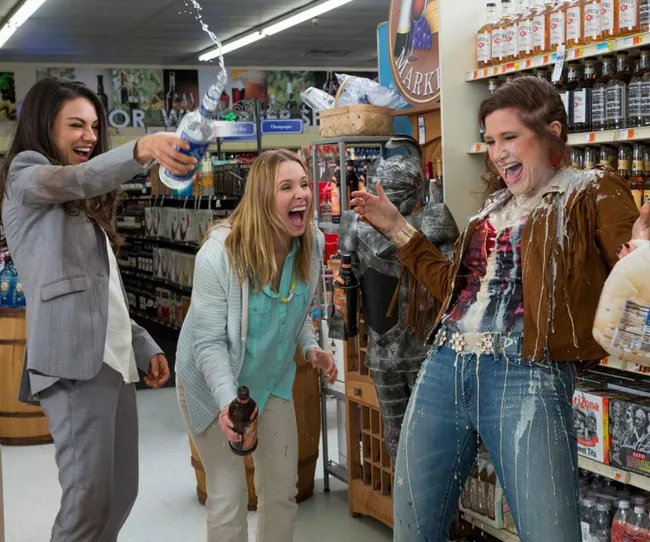 Three women laughing in a grocery store, with one pouring a drink on another, creating a playful mess.