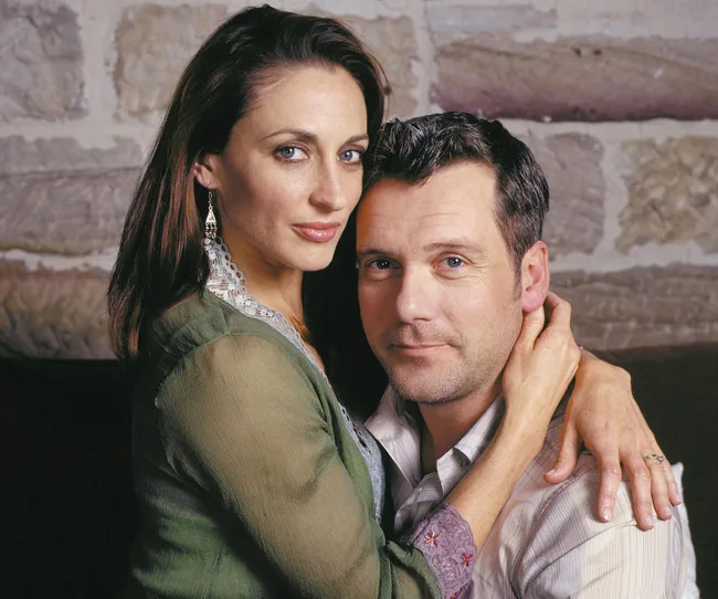 A woman in a green top embraces a man in a light shirt, both looking at the camera, against a stone wall background.