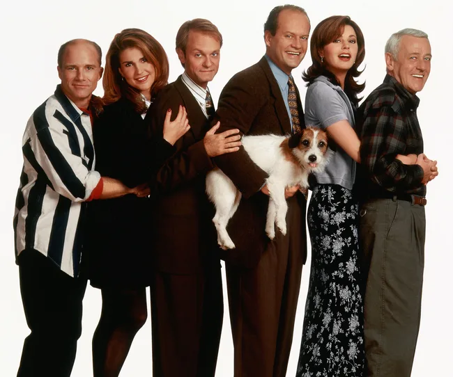 Group cast photo from "Frasier" with six people and a dog in a line, smiling, against a white background.