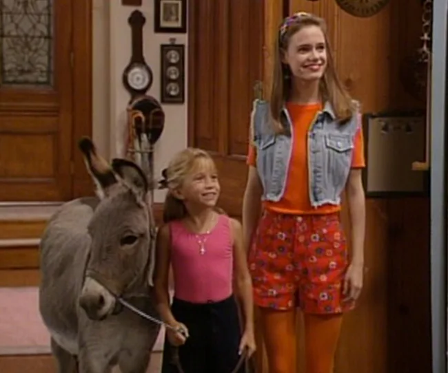 Young girl and older girl stand smiling indoors with a donkey.