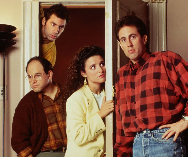Four people stand by a doorway, three looking outwards and one peering over the door, in a scene reminiscent of "Seinfeld."