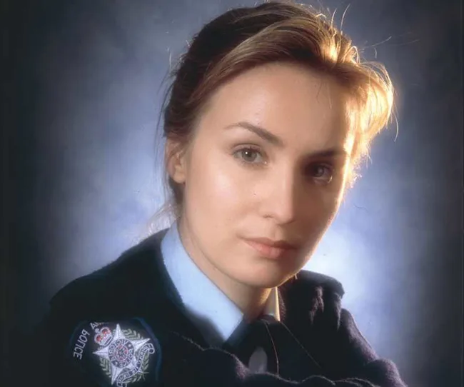A woman in a police uniform poses with a serious expression, set against a dark, blurred background.