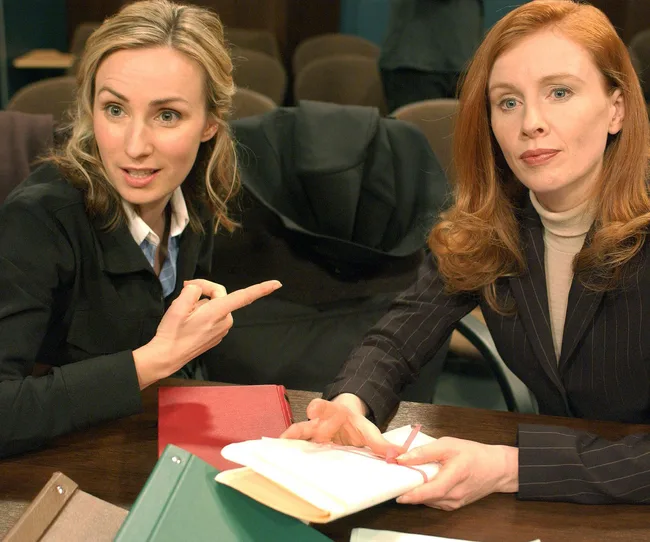 Two women sitting at a table with folders. One gestures while the other holds a pen and notebook, appearing in conversation.