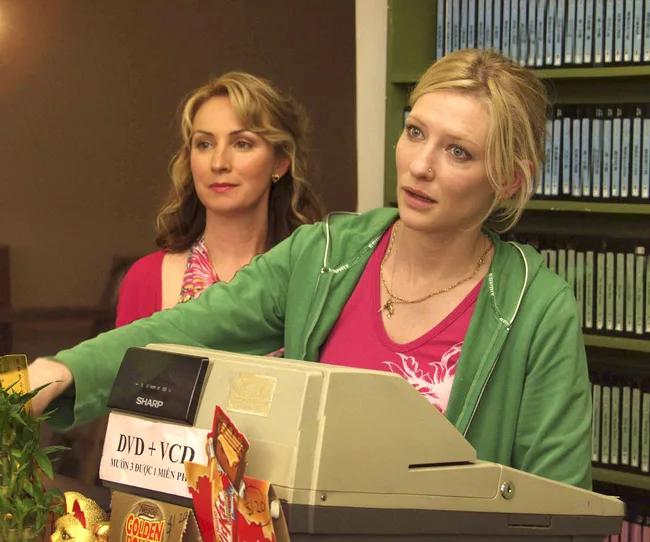 Two women stand behind a countertop with a cash register; one in a green hoodie and the other in a colorful scarf.