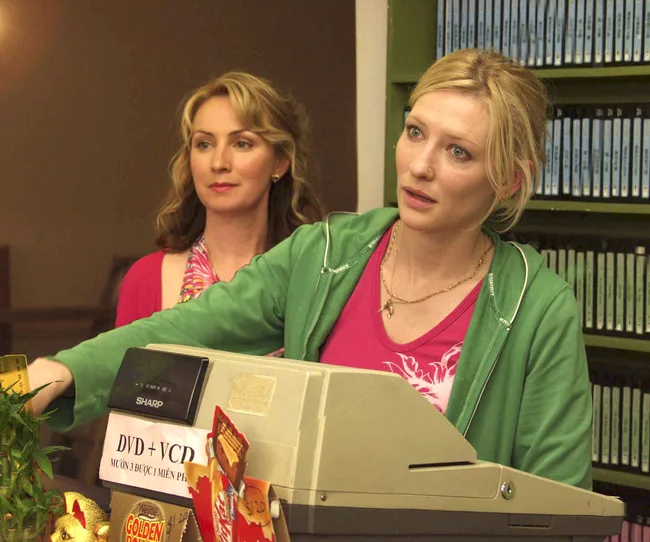 Two women stand behind a countertop with a cash register; one in a green hoodie and the other in a colorful scarf.