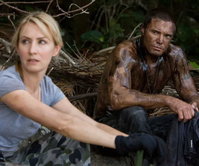 Two individuals in the jungle, one covered in mud, both appearing focused and contemplative.