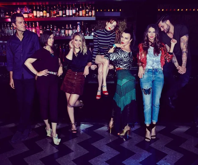 Group of seven people posing stylishly at a nightclub bar, wearing diverse outfits, smiling and interacting.