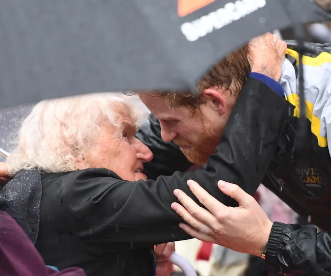 Prince Harry hugging an elderly woman under an umbrella in the rain in Sydney.