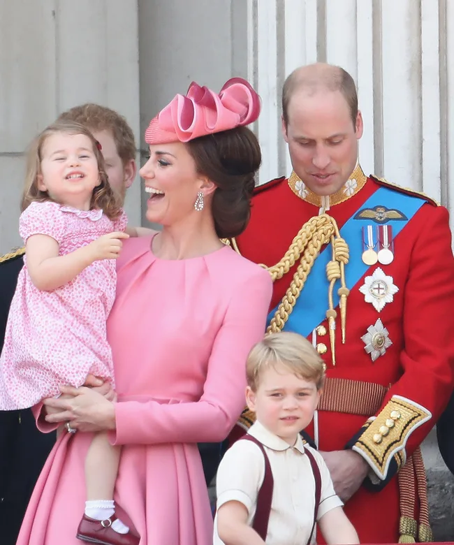 A family on a royal balcony, with a woman holding a smiling child, another child in front, and a man in uniform behind them.