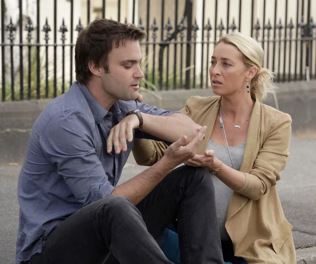 Man and woman sitting on a street; woman appears concerned, holding man's hand.