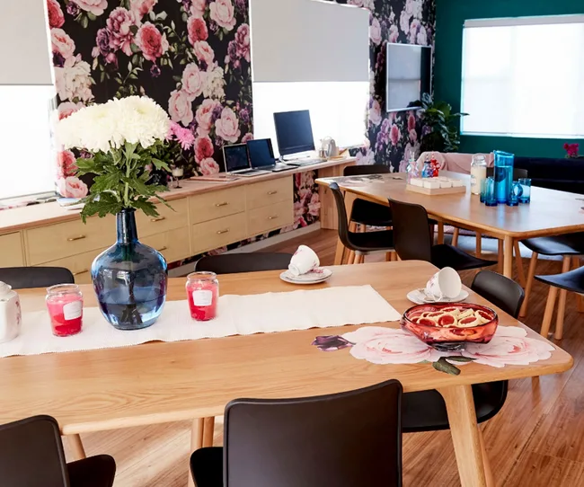 Floral-themed dining area with wooden tables, floral wallpaper, blue vase with flowers, and tea set.