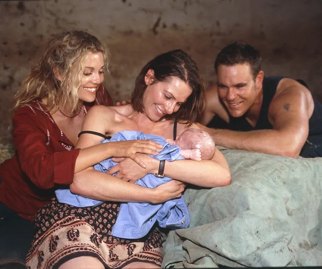 A joyful group of three adults smiles at a newborn baby wrapped in a blue blanket.