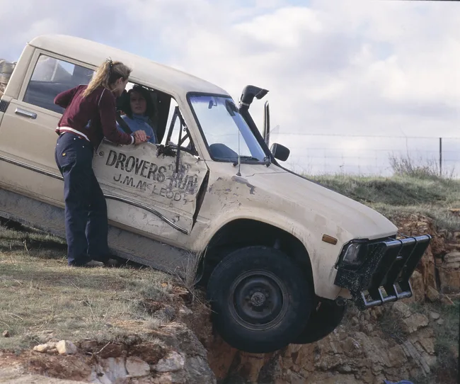 A woman in a car leaning over a cliff edge is assisted by another woman standing outside, by a vehicle labeled "Drovers Run."