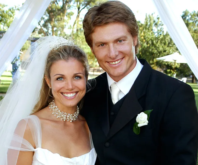 A bride and groom smiling at their outdoor wedding, with trees and a white arch in the background.