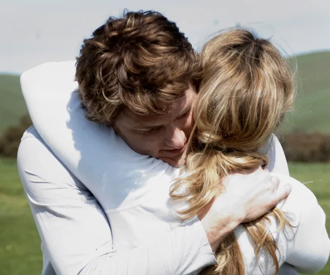 A couple embraces warmly in a grassy landscape, both wearing light-colored clothing.