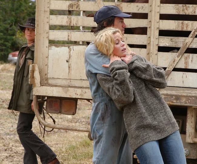 A woman in a gray sweater is held by a man in blue overalls. Another person in a hat watches near a wooden truck.