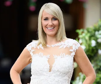 A woman in a white lace dress smiles outdoors, possibly at a garden party or wedding setting.